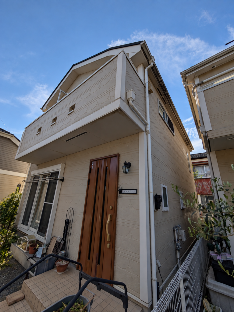 海老名市の戸建て住宅の屋根・外壁リフォーム前の外観。ベージュ系のサイディング外壁とバルコニー、木目調の玄関ドアが見える様子。